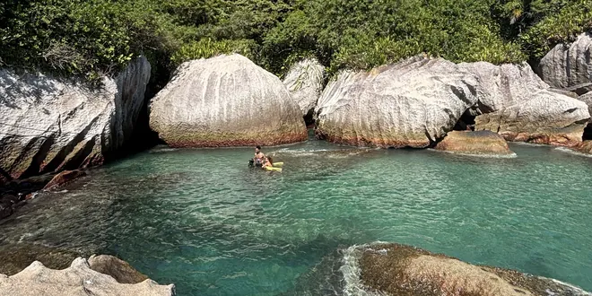 Piscina natural entre os costões, com uma pessoa boiando na água turquesa.